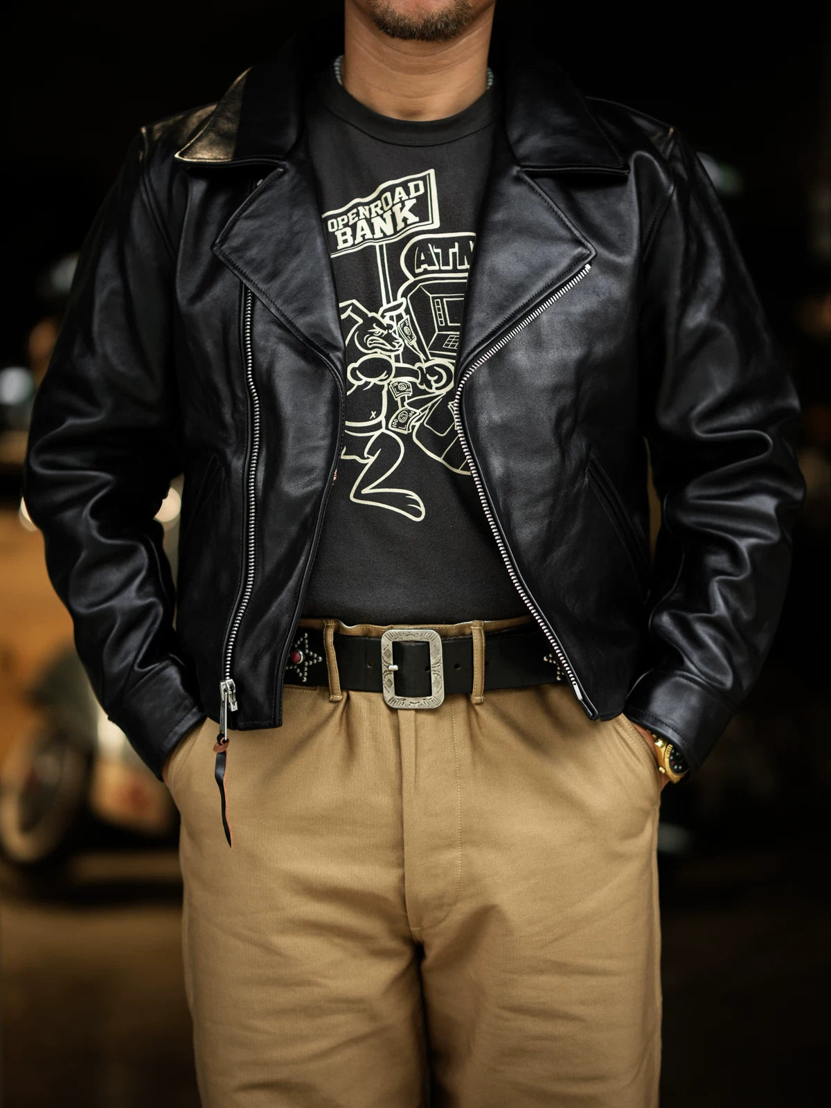 Model wearing black tea-core horsehide double-breasted aviator jacket open over graphic tee and khaki trousers, hands in pockets