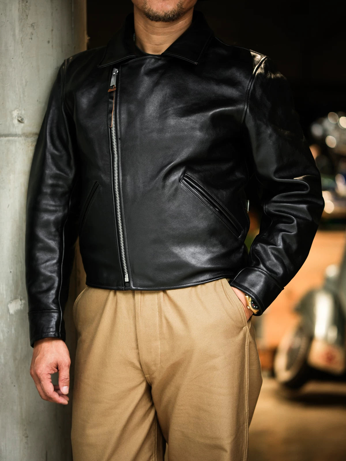 Black horsehide aviator jacket zipped to stand collar, model leaning against concrete pillar