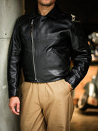 Black horsehide aviator jacket zipped to stand collar, model leaning against concrete pillar